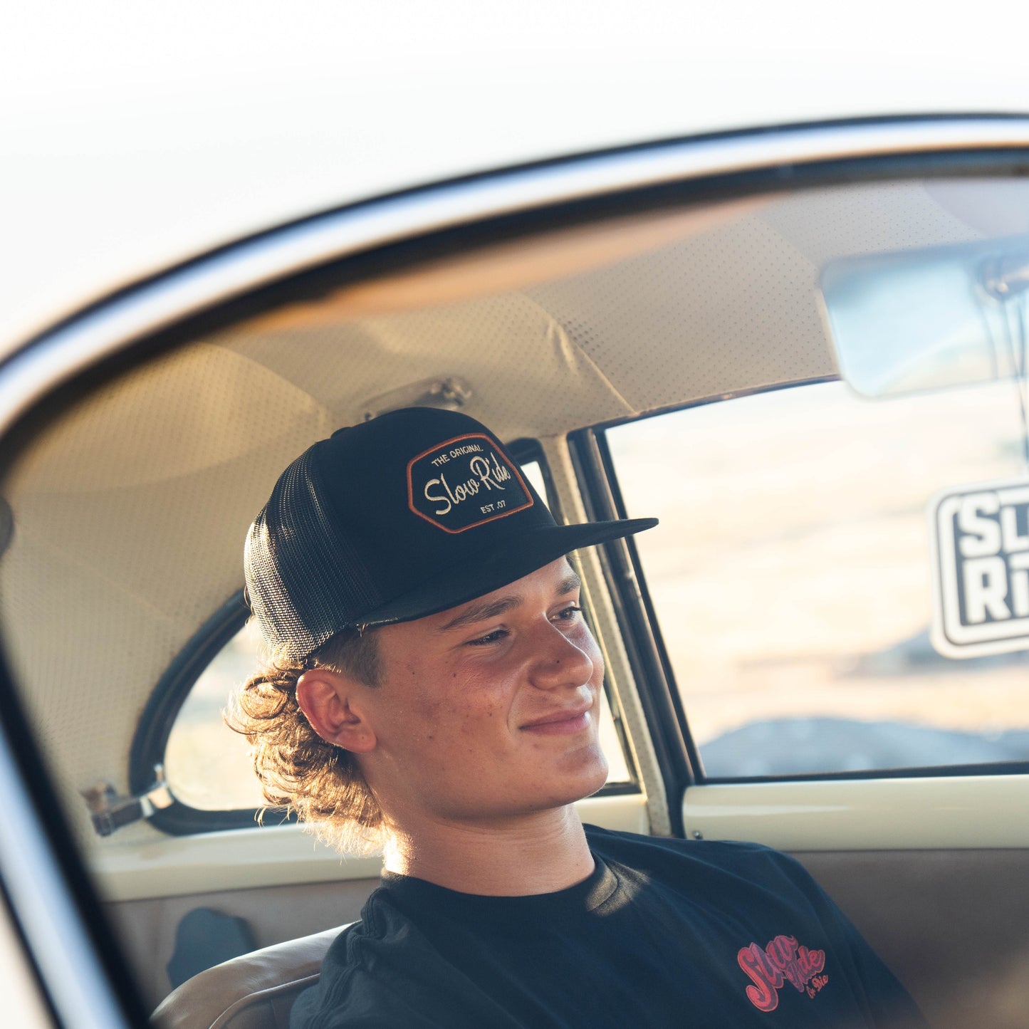 Patch Trucker Hat (Black) - Slow Ride- Black mesh trucker hat featuring a 'Slow Ride Garage' patch with 'The Original Slow Ride' and 'Est. 07' text in white and orange embroidery. Perfect for Off-Roading and Off-Road Racing enthusiasts, this hat pairs well with a slow ride t-shirt or shirt and celebrates the vintage Volkswagen culture, including the iconic Volkswagen bus, Volkswagen bug, and Baja bug. Ideal for those who embrace the Garage and Racing lifestyle and live by mottos like 'Enjoy Getting There,'